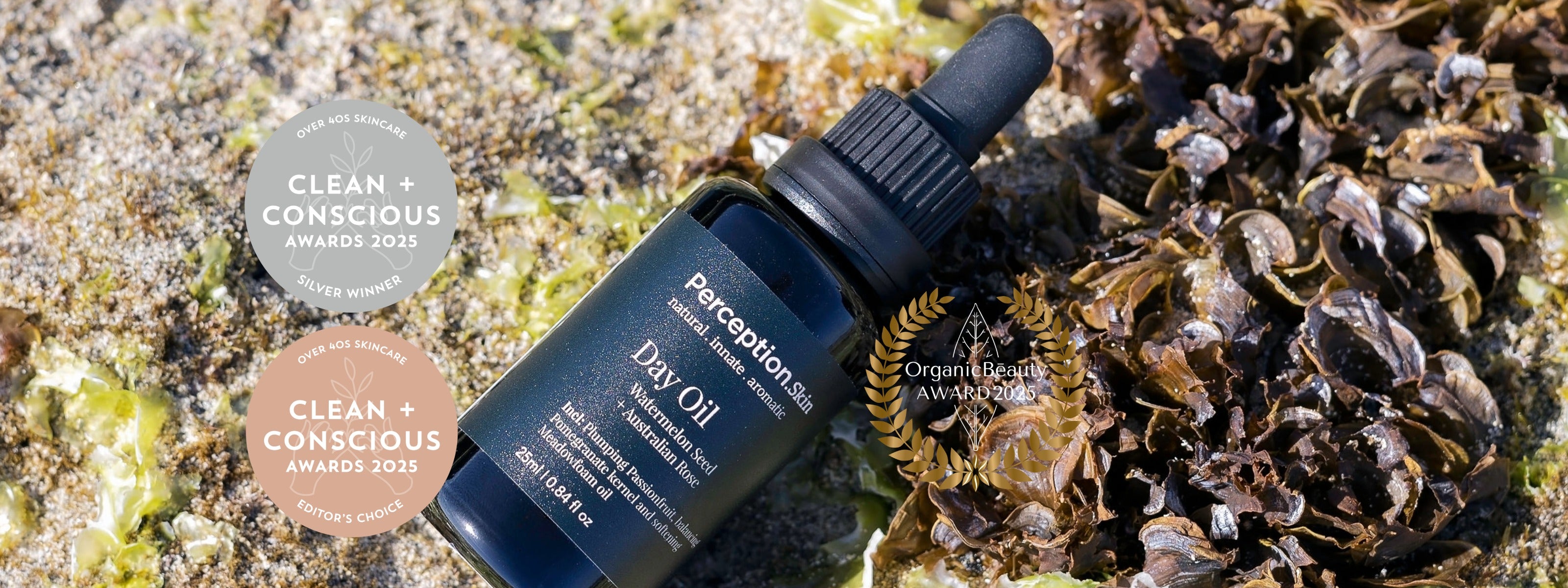 A dark blue glass dropper bottle labeled Perception Skin Day Oil placed on a natural sandy and seaweed background, with two circular award badges on the left.