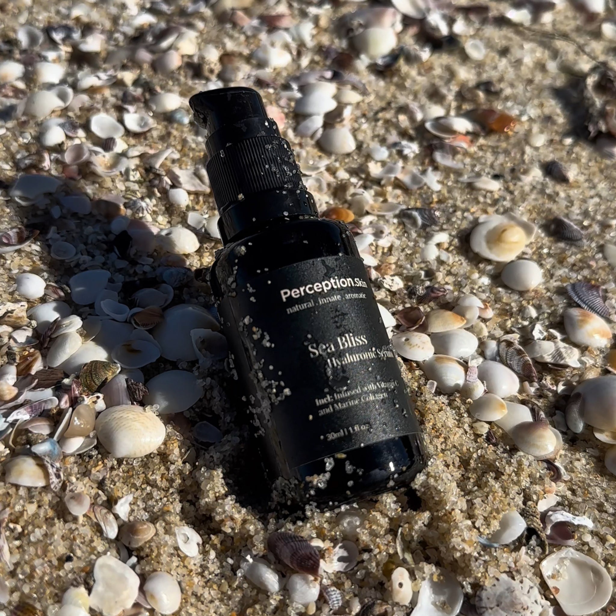 Perception Skin's Sea Bliss Hyaluronic Serum is placed on shells at the beach, product has sand on the bottle to show natural surrounding