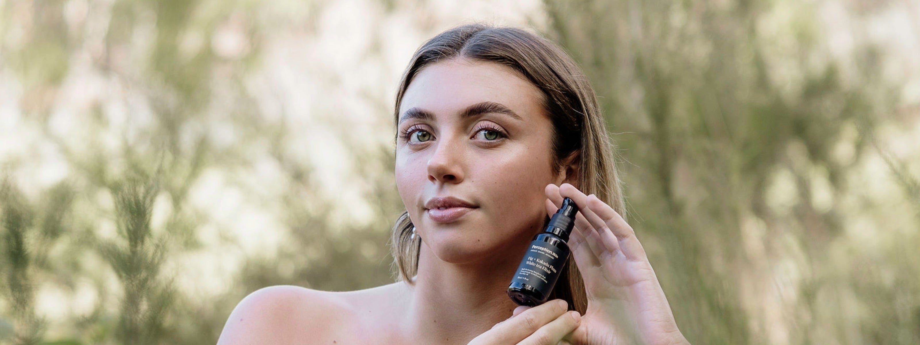 A woman outdoors holding Perception Skin near her face, with a softly blurred natural background.