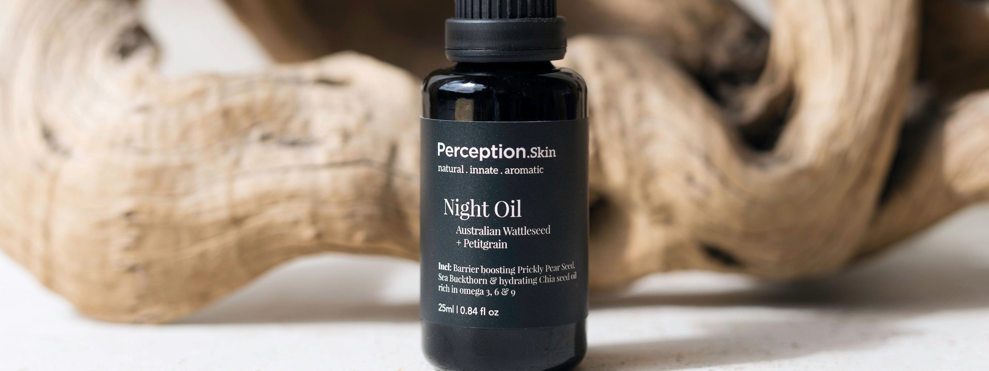 A small black bottle of Perception Skincare Night Oil with a black cap, set against a blurred background of natural driftwood on a light surface.