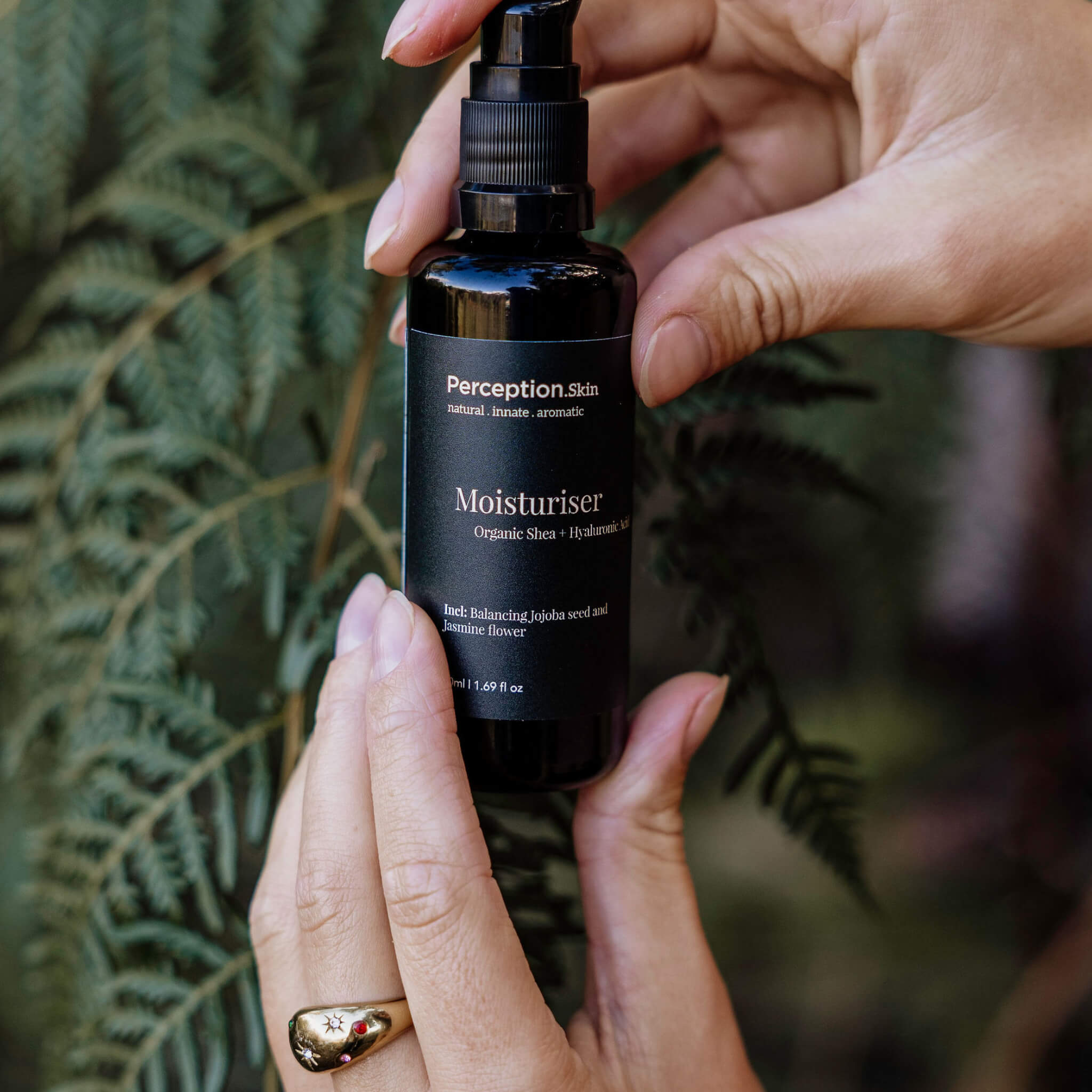 Hand holding a bottle of Perception Skincare Moisturizer with a blurred green leafy background