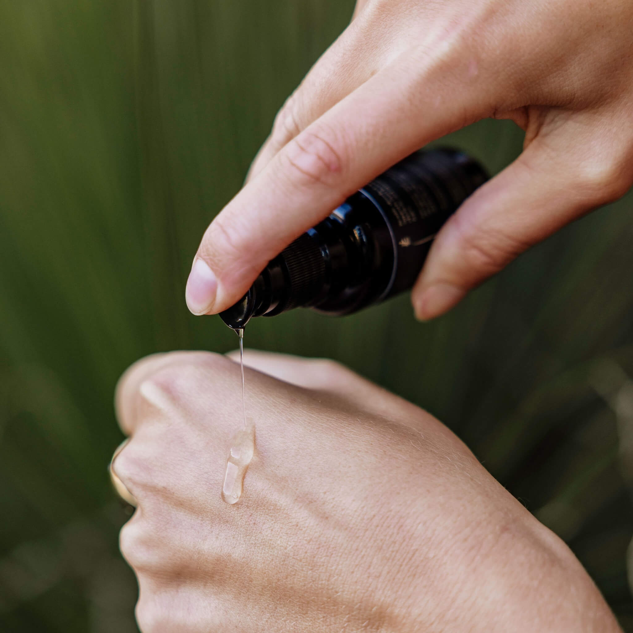 Hand holding a dropper bottle with a drop of liquid on another hand against a blurred green background